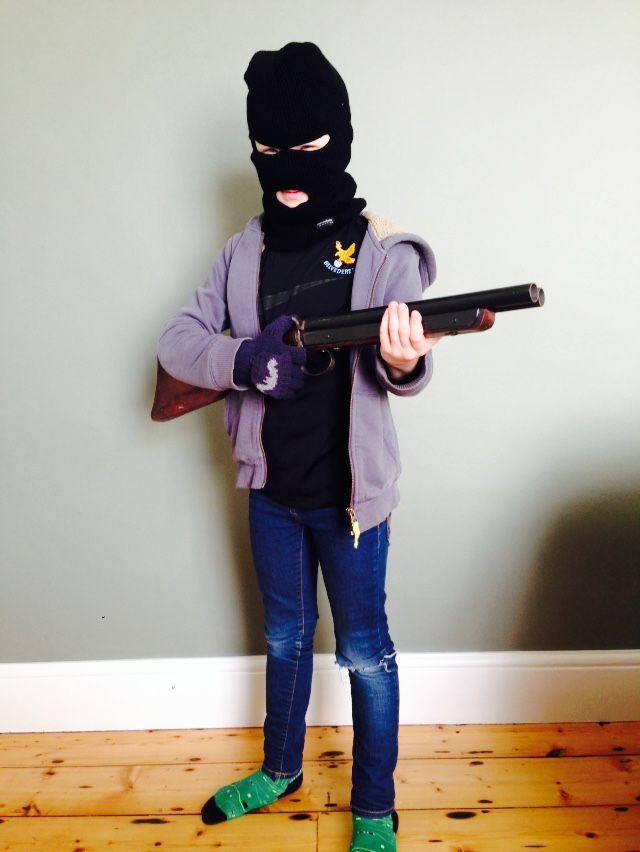 Person in a balaclava and jacket holding a rifle, standing indoors on a wooden floor.