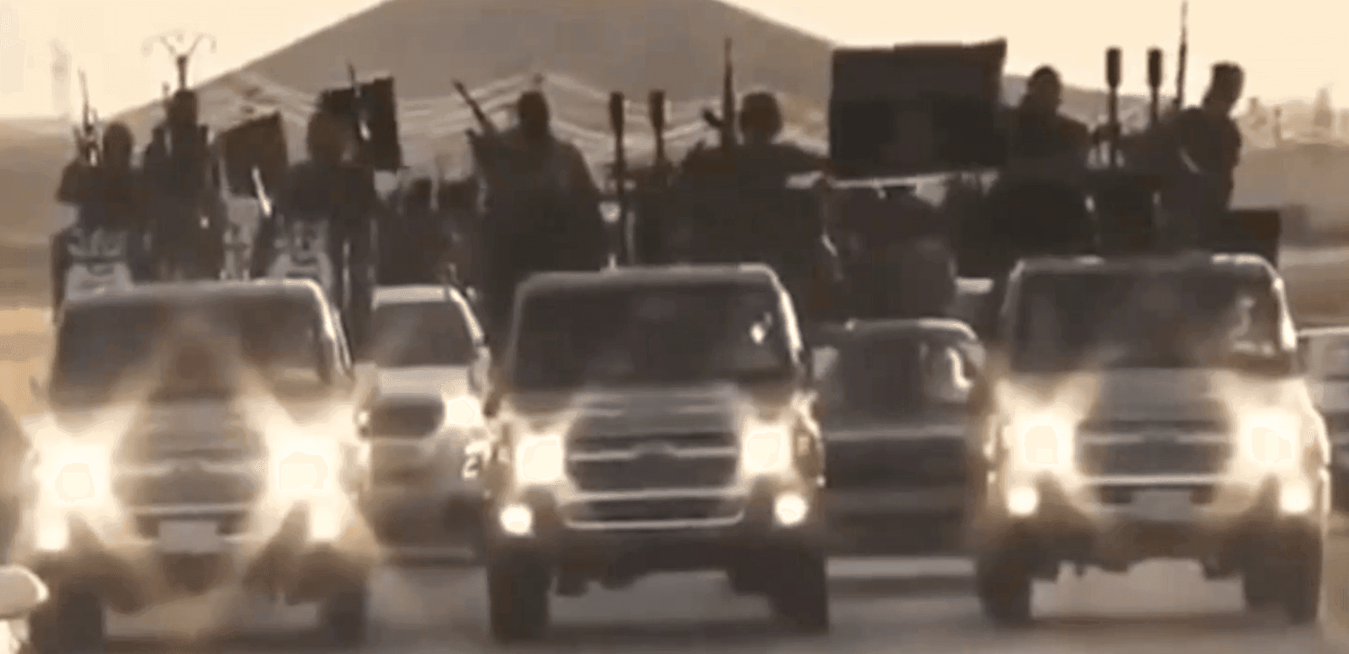 Convoy of vehicles with armed individuals driving on a road during dusk, headlights illuminating.