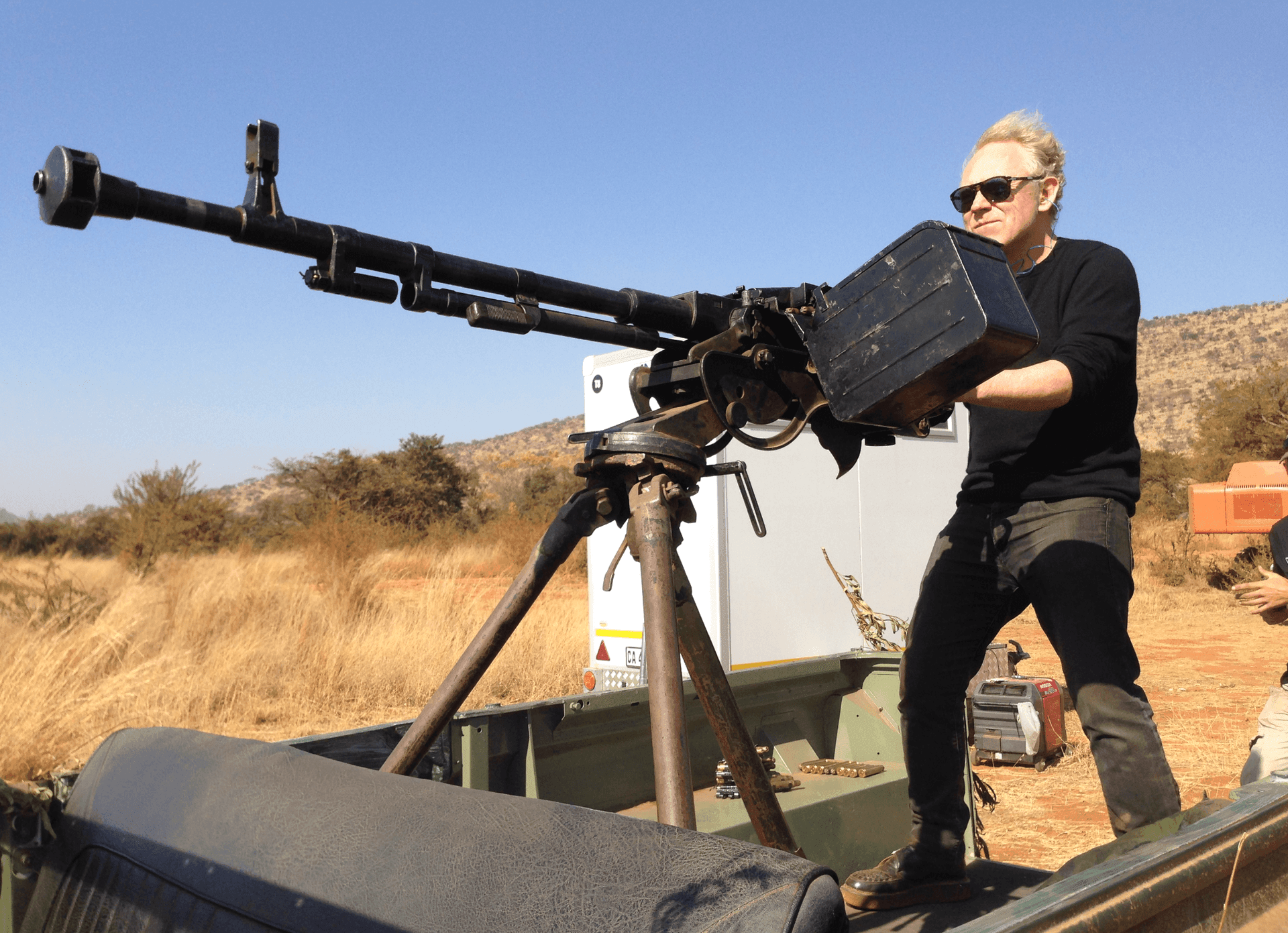 Person standing in a vehicle bed handling a mounted heavy machine gun in a dry, open landscape.